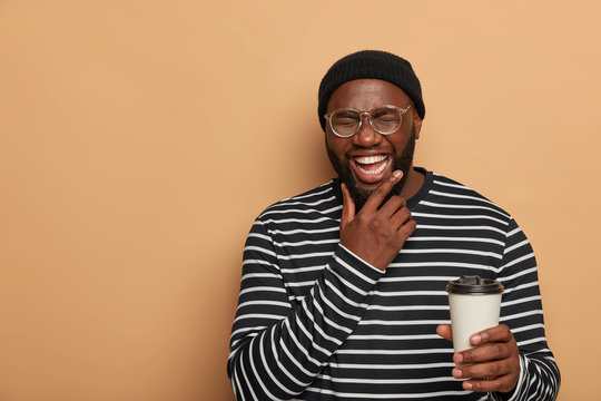 People, lifestyle, emotions concept. Joyful black man holds chin, smiles sincerely, drinks takeout coffee, dressed in stylish clothes, stands against beige background with copy space on left side