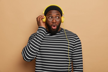 Indoor shot of emotional shocked bearded man listens modern music in headphones, looks with stupefied expression, surprised sound disappeared, wears black and white striped jumper, poses indoor