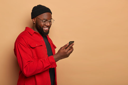 Profile shot of stylish dark skinned hipster in fashionable red shirt and black hat, holds smartphone and types email, gazes at display with happy smile, poses over beige background with empty space
