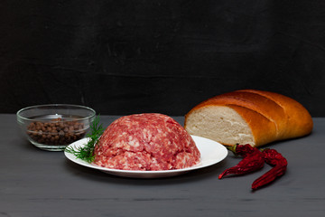 Minced meat on a white plate, dark background. Black pepper, chili pepper, onion, dill. Cutlets in Russian