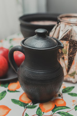 a jug of black ceramic stands on a beautiful tablecloth next to a plate with apples