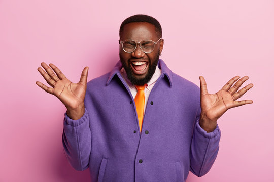 Portrait of overjoyed male boss raises palms, feels glad and satisfied, gets wonderful news, laughs from joy, has dark healthy skin, wears purple coat, poses in sudio against rosy background