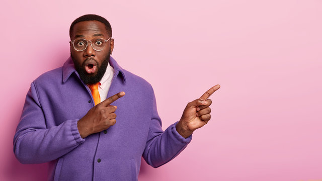 Stunned black bearded man with thick stubble shows something surprising at copy space, opens mouth with shock, wears fashionable clothes, stands over pink bacground, cannot believe his eyes.