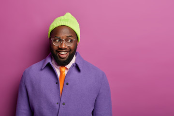Studio shot of happy black Afro American man looks gladfully aside, wears stylish purple jacket, hat and optical glasses, expresses sincere emotions, models against bright background, blank space