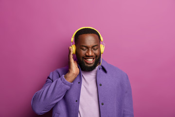 Obraz premium Smiling black man enjoys good sound in headphones, made new playlist, listens favourite music during spare time, dressed in purple jacket, shows white teeth. People, lifestyle, hobby concept