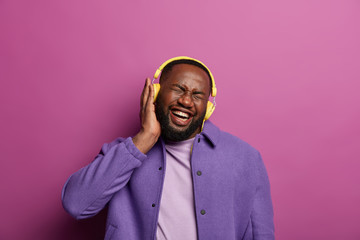 Obraz premium Enegrized overjoyed hipster listens loud music in yellow headphones, laughs and closes eyes, forgets about all problems, feels upbeat during listening nice popular songs in playlist, has spare time