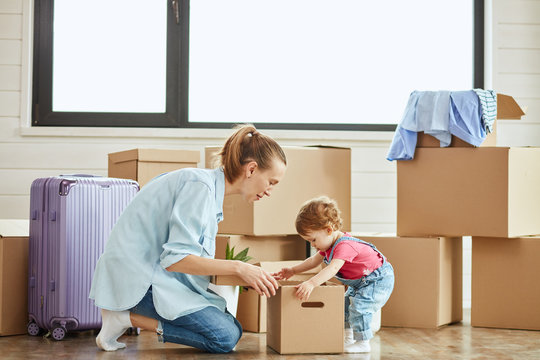 In center photo little child looks in box. Mother blonde woman wear blue shirt, jeans and white socks smile and look into box. Background moving boxes and suit