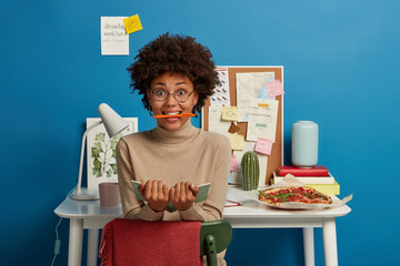 Surprised Afro American woman has fun while working, keeps pen in mouth, holds opened notebook for...