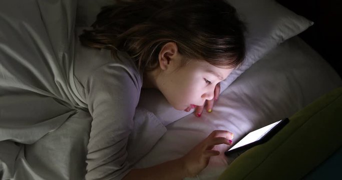 face lit of six years old girl typing on digital display of smartphone lying on bed at night  