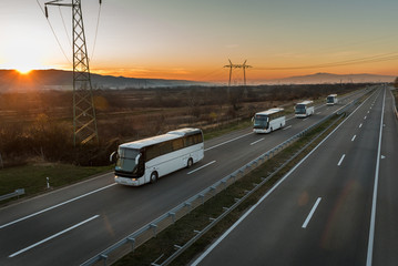 Caravan or convoy of Four white buses in line traveling on a country highway under amazing orange...