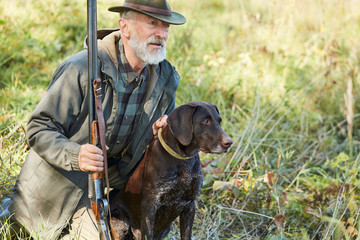 Caucasian mature man with gun and dog sit searching prey. Bearded man in hunting clothes. Autumn