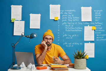 Concentrated tired schoolboy in yellow clothes, prepares homework project, holds pen, eats fast...