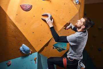 Fototapeta na wymiar Side view of professional rock-climber without forearm training indoors at colorful climbing wall in bouldering gym, prepairing to oncoming ascend, working out most complicated problems.