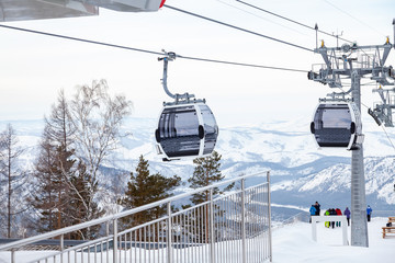 Obraz premium Landing place to a gondola cableway booth suspended on a cable where sits people with skis and snowboards on a background of mountains, trees with snow on winter sunset. Ski resorts and snowboarding.