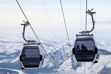 Cabin of a gondola cableway suspended on a rope where sits people with skis and snowboards high in the Altai mountains with snow and blue sky on winter sunset. Ski resorts and snowboarding.