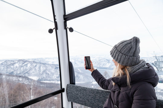 A young beautiful blonde girl in winter clothes and a gray knitted hat sits in the gondola lift cabin in the mining resort and takes photos of the mountains and landscape Altai.