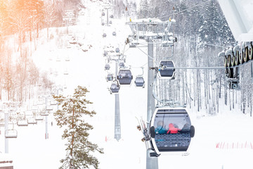 A gondola cableway with a booth suspended on a cable in which sits people with skis and snowboards on a background of mountains, trees with snow on winter sunset. Ski resorts and snowboarding.