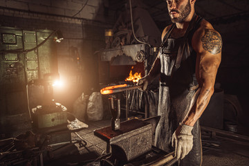 forger shop owner at work, stand heating red metal wearing gloves and apron isolated in dark space