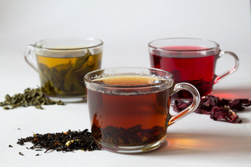 Black hot tea with dry leaves.Three glass cups with black, red and green tea with dry leaves on a white background. Red hot Hibiscus tea with dry flowers. Healthy lifestyle. Green hot tea. Hot drinks.