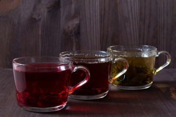 Three glass cups with black, red and green tea on a dark wooden background. Red hot Hibiscus tea. Black hot tea. Healthy lifestyle. Hot drinks. Green hot tea on wooden table.
