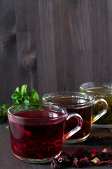 Healthy lifestyle. Hot drinks. Three glass cups with black, red hibiscus and green herbal hot tea with dry leaves and flowers with fresh mint on a dark wooden background. Vertical orientation.