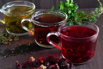 Three glass cups with black, red hibiscus and green herbal hot tea with dry leaves and flowers with fresh leaves of mint on a dark wooden background. Healthy lifestyle. Hot drinks.