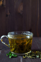 Glass cup of green herbal hot tea with dry leaves with fresh green leaves of mint on a dark wooden background. Hot drinks. Vertical orientation. copy space. Healthy lifestyle.