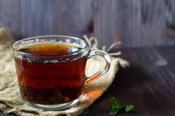 Cup of black tea with fresh green leaves of mint on a wooden table. Healthy lifestyle. Glass cup of black hot tea with sackcloth on a dark wooden background. Hot drinks.