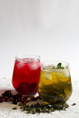 Fototapeta na wymiar Glasses of iced green herbal tea and red hibiscus tea with dry leaves and flowers on a white table. Green iced tea and fresh green leaves of mint on white background. Healthy lifestyle. Cold drinks.
