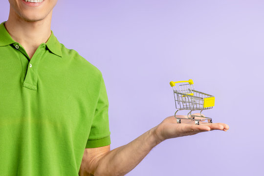 Portrait Of Cropped Young Man In Green T-shirt Holding Small Toy Truck For Shopping From Hypermarket, Isolated Over Purple Background