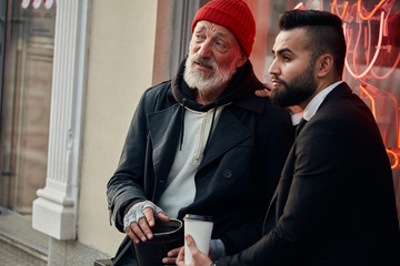 Rich man in tuxedo and homeless person in dirty clothes sitting and talking in the street. Poverty, help