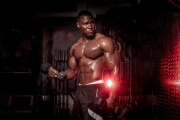 Muscular African American, black shirtless , sweaty male bodybuilding athlete does barbell curls in a dark grungy gym with dramatic lighting flare