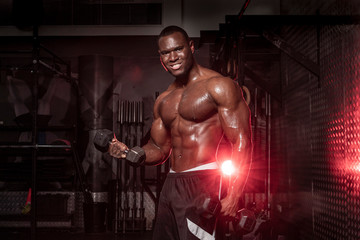 Fototapeta na wymiar Muscular African American, black shirtless , sweaty male bodybuilding athlete does barbell curls in a dark grungy gym with dramatic lighting flare