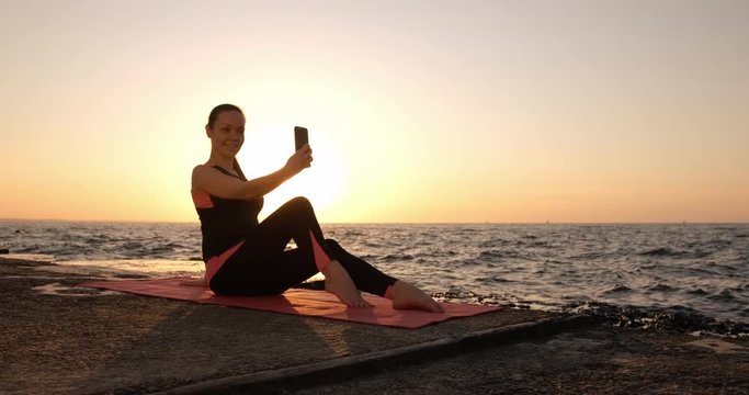 A young slender girl in a fitness suit sits on a pier by the ocean at dawn and takes pictures of herself on a smartphone.