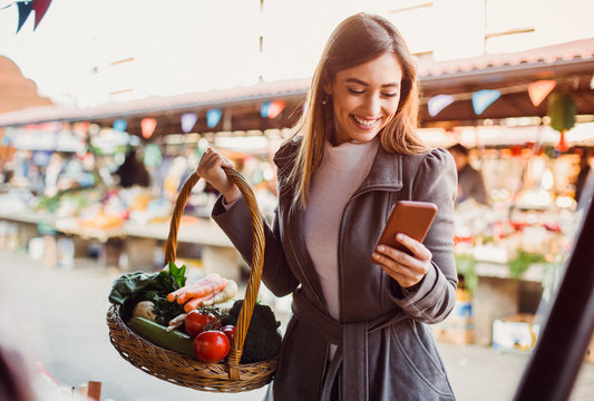 Beautiful young brunette text messaging on her smart-phone while she's doing shopping at farmers' market.