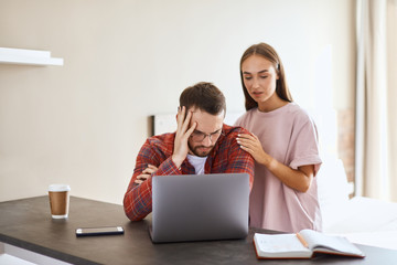 Serious, upset husband wears round spectacles, stares at laptop computer, sitting at table in bedroom, holds head with dissatisfied expression, portrait, indoor shot