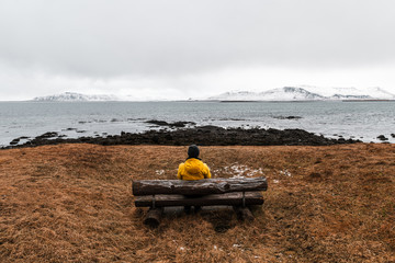 Lonely traveler man in yellow rain jacket  is relaxing at nice view of sea and mountain with wooden bench in Reykjavik, Iceland.