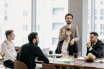 funny caucasian business colleagues sit on table and have conversation during free time at work, everyone in formal wear. one man grabbed the head of another. panoramic window in the background