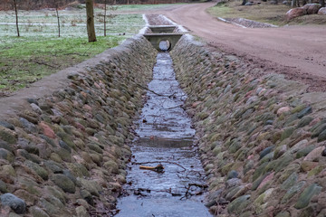 A ditch full of runoff water flowing along the road lined with stone in the countryside