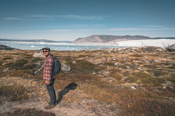 Obraz premium A young man traveler tourist standing in front of Eqip Sermia glacier called Eqi Glacier. Wall of ice in background. The concept of global warming and professional guides. On a sunny day with blue sky