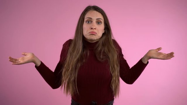 Portrait of attractive young long hair woman feeling perplexity and misunderstanding emotions posing isolated over studio pink background. Helpless woman throwing hands wide and shaking shoulders