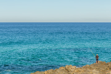 Fototapeta na wymiar Beautiful blue ocean, a single fisherman stands on rocks alone with small blue waves