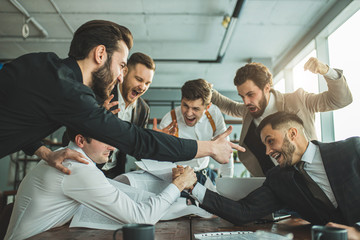 leisure time of happy business team at work in office, two bearded men in tuxedo playing arm wrestling, their colleagues support and cheering for them.