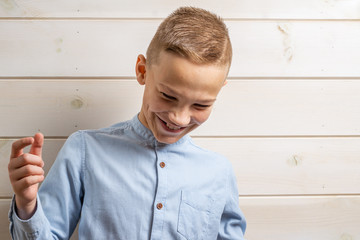 A boy of 10 years old in a blue shirt smiles on a light wooden background and makes various signs with his hands.