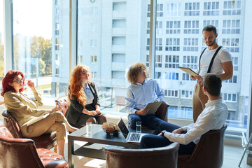 A group of caucasian people, 2 woman and 3 men coworking together at the openspace office. Having a conversation, sharing knowledge and skills. One man is reading his ideas and data.