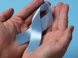 Close-up male hands holding blue prostate cancer awareness ribbon on a bright blue background....