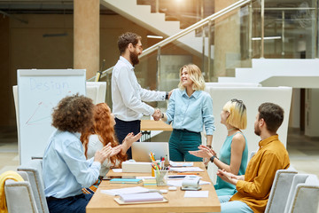 Nice, kind girl dressed in casual jeans and light blue shirt, looks happily at satisfied bearded team leader with tender smile, gives friendly handshake , designers modern office concept
