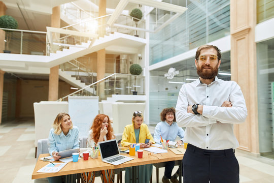 Self-assured satisfied man in stylish round glasses, wears formal clothes, keeps hands folded, expresses proudness, looks straight at camera, posing on background of colleagues