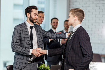 boss in stylish tuxedo congratulate best worker of company with new job position, guy happy to be part of successful business team, boss and man shaking hands while colleagues stand in background