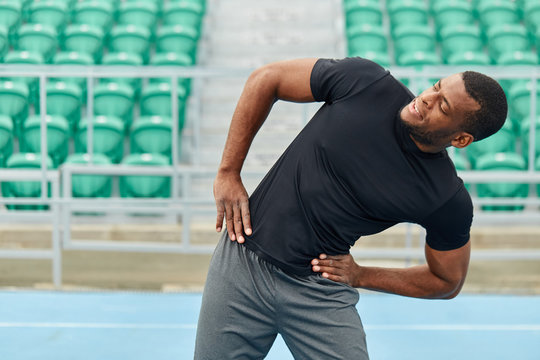 african cheerful sportsman holding arms on the hips doing side bends outdoors. close up photo, colorful seats in the background of the photo. fitness, sporty lifestyle